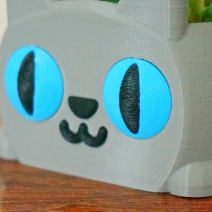 Pode incluir: Um vaso de gato cinza impresso em 3D com olhos azuis e um nariz e boca pretos. O vaso est&aacute; sentado em uma superf&iacute;cie de madeira.