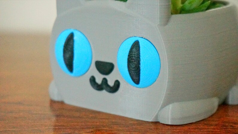 Pode incluir: Um vaso de gato cinza impresso em 3D com olhos azuis e um nariz e boca pretos. O vaso est&aacute; sentado em uma superf&iacute;cie de madeira.