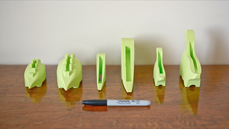 May include: A set of six lime green 3D printed phone stands of varying sizes. The stands are arranged in a row on a wooden surface.