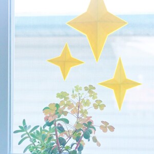 May include: A white ceramic planter shaped like a cartoon character with red cheeks and black eyes. The planter is filled with a variety of green succulents and is sitting on a wooden tray. Three yellow paper stars are adhered to the window behind the planter.