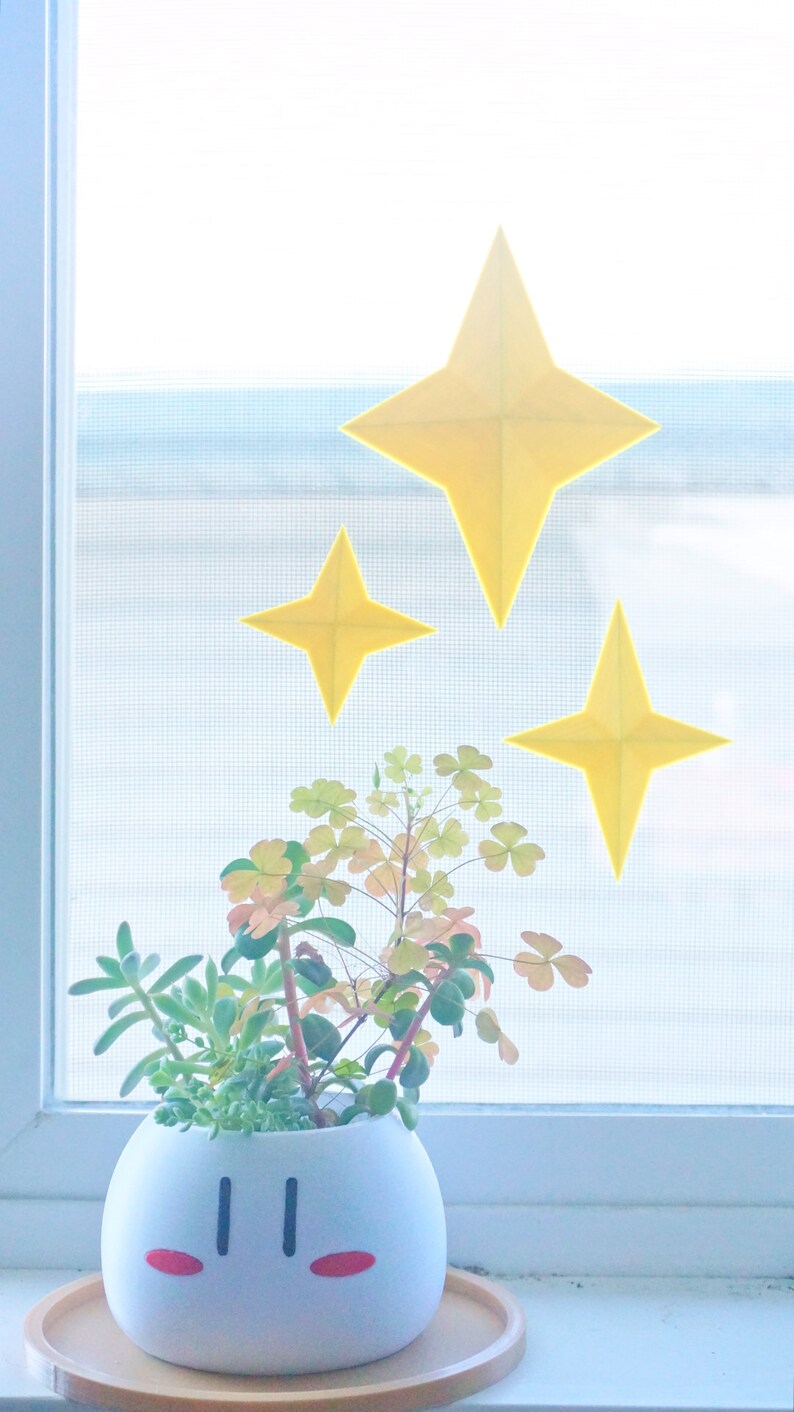 May include: A white ceramic planter shaped like a cartoon character with red cheeks and black eyes. The planter is filled with a variety of green succulents and is sitting on a wooden tray. Three yellow paper stars are adhered to the window behind the planter.
