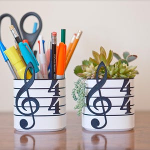 May include: Two white cylindrical containers with musical note designs. One holds pens, pencils, and scissors. The other contains a small succulent plant. The musical notes are black, and the containers are on a wooden surface.