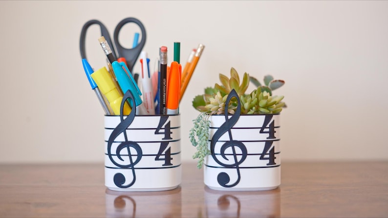 May include: Two white cylindrical containers with musical note designs. One holds pens, pencils, and scissors. The other contains a small succulent plant. The musical notes are black, and the containers are on a wooden surface.