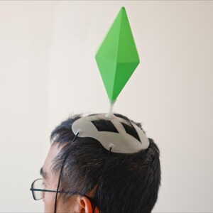 May include: A person wearing a white and black headpiece with a large green diamond shape attached. The headpiece is secured with black strings. The person is wearing glasses and is against a white background.