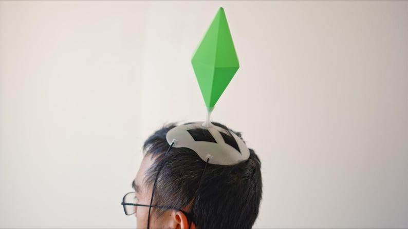 May include: A person wearing a white and black headpiece with a large green diamond shape attached. The headpiece is secured with black strings. The person is wearing glasses and is against a white background.
