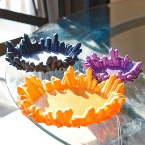 May include: Three decorative crystal-shaped dishes in blue, purple, and orange. The dishes are made of a material that resembles crystal and are arranged on a glass table.