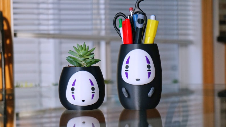 May include: Two black desk organizers with a white and purple face design. One holds a small succulent plant, the other holds pens, pencils, and scissors. The organizers are on a reflective surface.