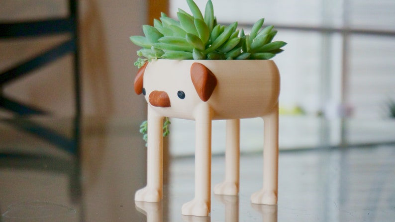 May include: A beige ceramic planter shaped like a pug with brown ears and a small succulent plant growing inside. The planter has four legs and is standing on a table.