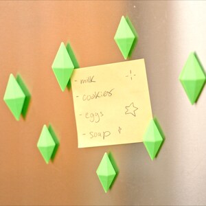 May include: A yellow sticky note with a handwritten grocery list on a silver refrigerator. The list includes milk, cookies, eggs, and soap. The note is held in place by six green diamond-shaped magnets.