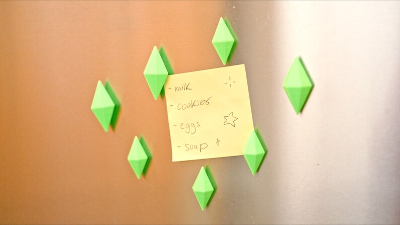 May include: A yellow sticky note with a handwritten grocery list on a silver refrigerator. The list includes milk, cookies, eggs, and soap. The note is held in place by six green diamond-shaped magnets.