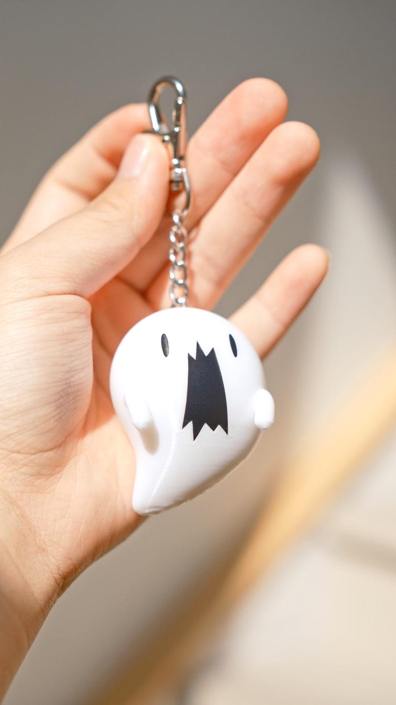 May include: A white ghost-shaped keychain with a black mouth and eyes. The keychain is attached to a silver chain and clasp. The ghost has a rounded body and small arms. The background is blurred.
