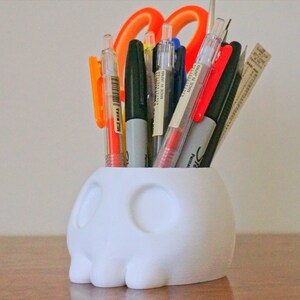 May include: White skull-shaped desk organizer with a variety of pens and a pair of orange scissors inside.