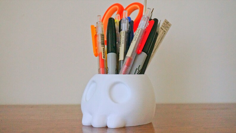May include: White skull-shaped desk organizer with a variety of pens and a pair of orange scissors inside.