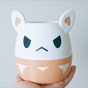 May include: White ceramic planter with a cartoon face, featuring black eyes, a frown, and two small pink hearts on the ears. The planter has a light brown band around the bottom.