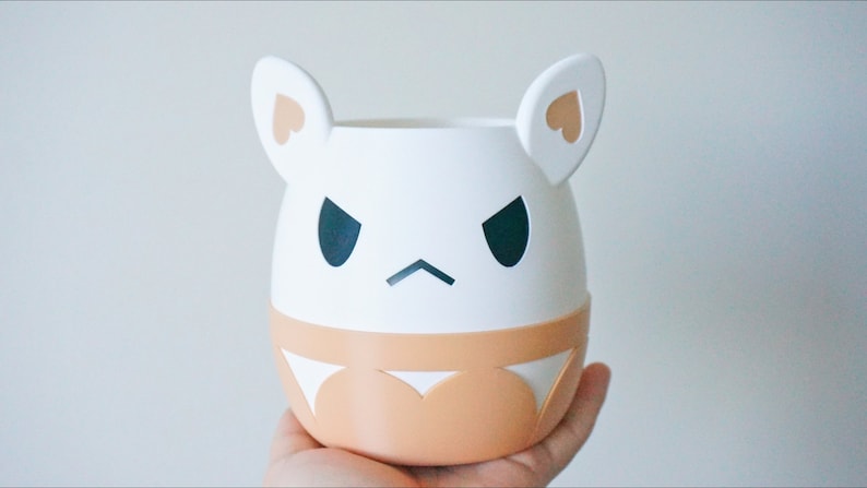May include: White ceramic planter with a cartoon face, featuring black eyes, a frown, and two small pink hearts on the ears. The planter has a light brown band around the bottom.
