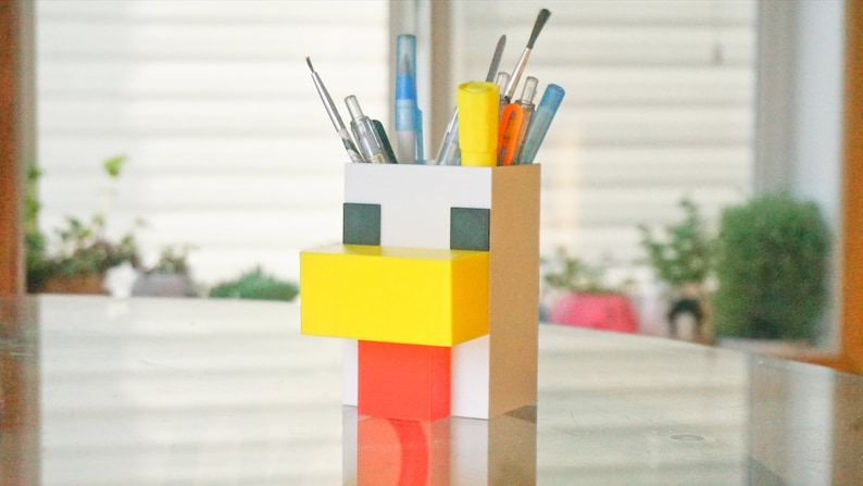 May include: A white, yellow, and red cardboard pencil holder shaped like a Minecraft chicken. The holder has a square design and features a yellow beak, red wattle, and black eyes.
