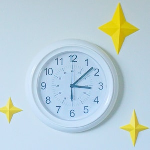 May include: A white analog clock with black hands and numbers on a white wall with three yellow paper stars.