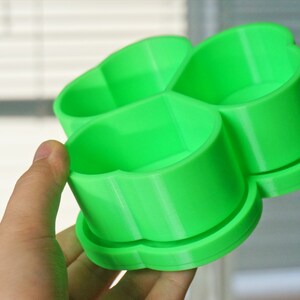 May include: A bright green, four-sectioned container, held in a hand. The container has a clover-like shape, with each section appearing to be a small cup. The object is made of plastic and has a smooth surface.