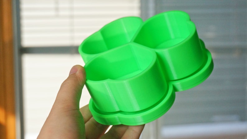 May include: A bright green, four-sectioned container, held in a hand. The container has a clover-like shape, with each section appearing to be a small cup. The object is made of plastic and has a smooth surface.