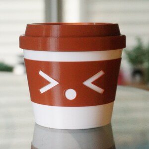 May include: A white and brown to-go coffee cup with a cartoon face on the side. The face has two white arrows pointing outwards and a white circle in the middle.