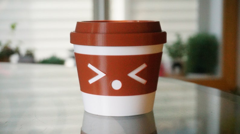 May include: A white and brown to-go coffee cup with a cartoon face on the side. The face has two white arrows pointing outwards and a white circle in the middle.