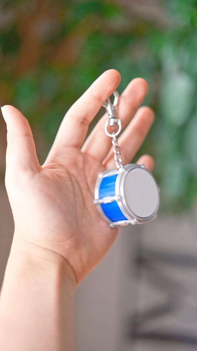 May include: A miniature drum keychain with a blue body and silver accents. The drum has a white surface and is attached to a silver chain and clasp. The keychain is held in a hand against a blurred background.