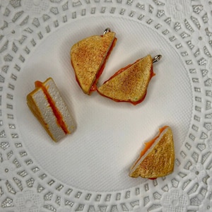 May include: Four miniature grilled cheese sandwiches with orange cheese on a white background. The sandwiches are made of polymer clay and are about the size of a quarter.