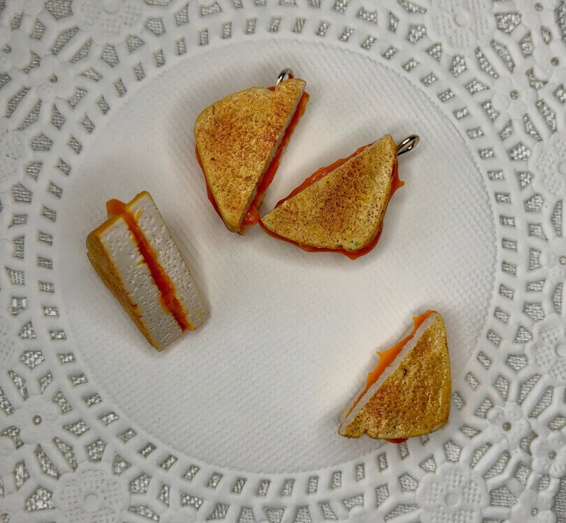 May include: Four miniature grilled cheese sandwiches with orange cheese on a white background. The sandwiches are made of polymer clay and are about the size of a quarter.