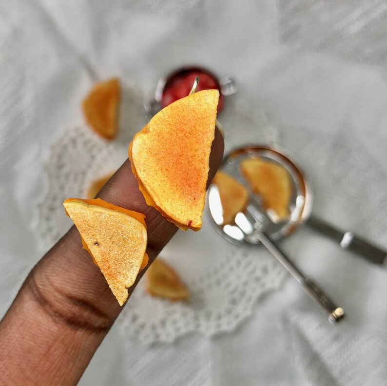 May include: Two small, orange-brown, grilled cheese sandwich earrings. The earrings are on a finger, and the background is white.