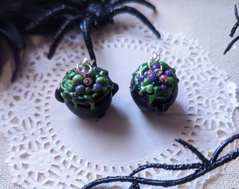 May include: A pair of miniature black cauldron earrings featuring a bubbling green potion mixture with small purple and green spheres and a single eyeball detail. Each cauldron is topped with a silver-toned metal jump ring for attachment.