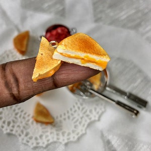 May include: A miniature grilled cheese sandwich with melted cheese inside, made of polymer clay. The sandwich is on a finger and there are other miniature grilled cheese sandwiches in the background.