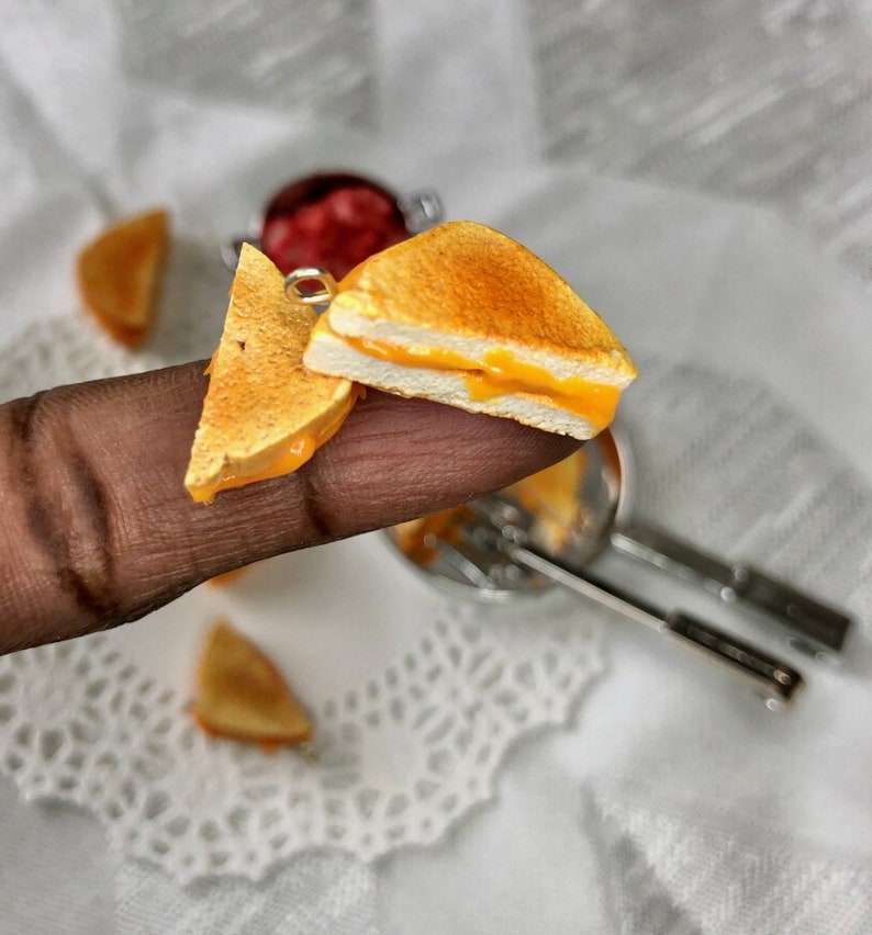 May include: A miniature grilled cheese sandwich with melted cheese inside, made of polymer clay. The sandwich is on a finger and there are other miniature grilled cheese sandwiches in the background.