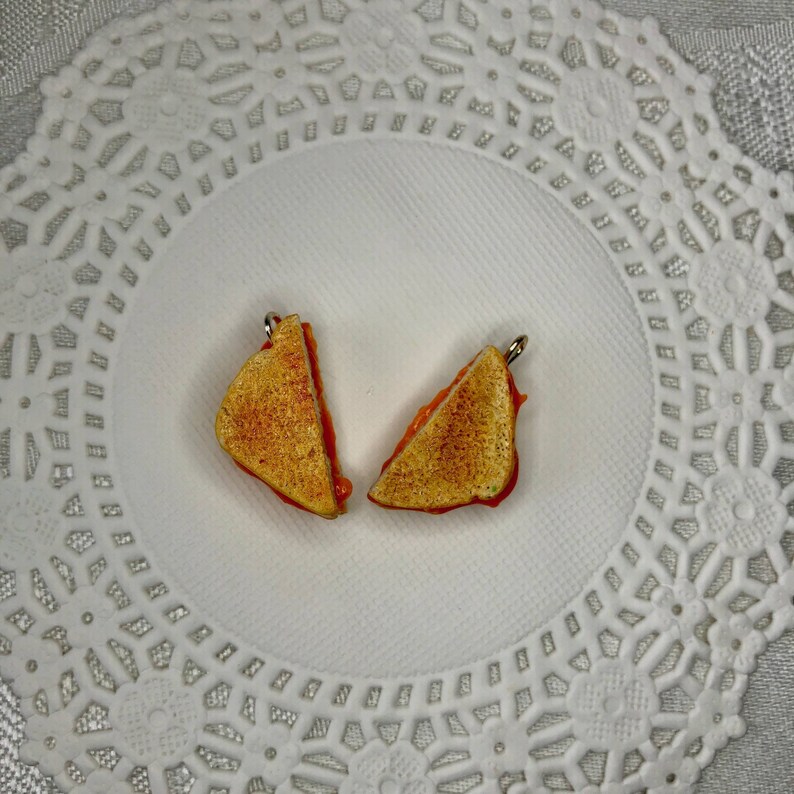 May include: Two miniature grilled cheese sandwiches with a red filling, each with a gold loop for hanging, on a white doily.