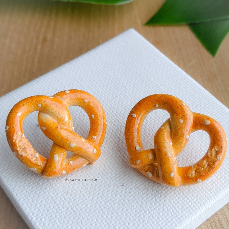 May include: A pair of pretzel-shaped earrings made of polymer clay. The earrings are a light orange color with white sprinkles on top. The earrings are on a white surface.