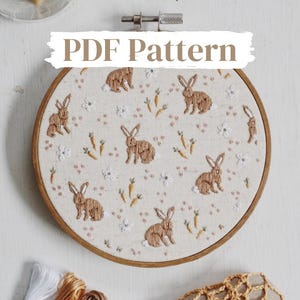 May include: A PDF pattern for embroidery featuring a design of brown bunnies and carrots on a white background. The pattern is displayed in a wooden embroidery hoop.