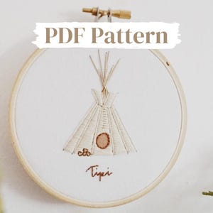 May include: A white embroidery hoop with a beige embroidered tipi design. The embroidery includes brown stitching and the word "Tipi" is stitched in brown thread below the tipi.