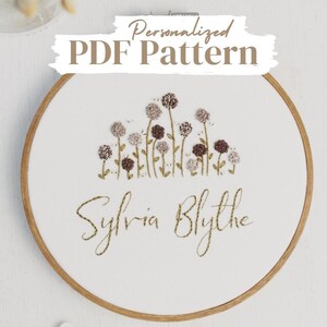 May include: A white embroidery hoop with a brown wooden frame. The hoop features a design of brown and white flowers with green stems and the name "Sylvia Blythe" embroidered in brown thread.