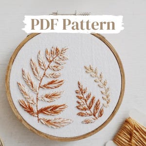 May include: A PDF pattern for embroidery featuring a design of three fern leaves in shades of brown and white on a white fabric background. The leaves are stitched in a hoop.
