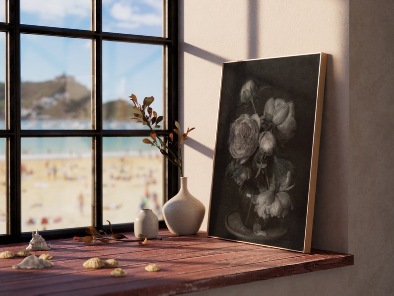 May include: A framed black and white print of a bouquet of peonies in a vase, sitting on a wooden windowsill with a view of a beach and a distant town. There are two small white ceramic vases and seashells on the windowsill.