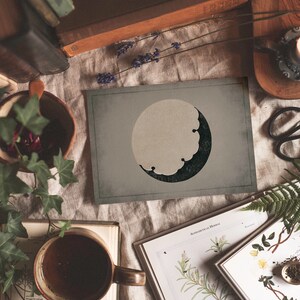 Puede incluir: Una impresi&oacute;n de estilo vintage de una luna creciente con un dise&ntilde;o verde oscuro y negro sobre un fondo gris claro. La impresi&oacute;n est&aacute; sobre una mesa con libros, plantas y otros art&iacute;culos decorativos.