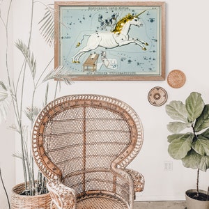 K&ouml;nnte beinhalten: Eine Vintage-Illustration des Sternbilds Einhorn, mit einem wei&szlig;en Einhorn mit goldenem Horn, vor einem blauen Hintergrund mit wei&szlig;en Sternen. Die Illustration ist in einem hellen Holzrahmen gerahmt und h&auml;ngt an einer wei&szlig;en Wand.