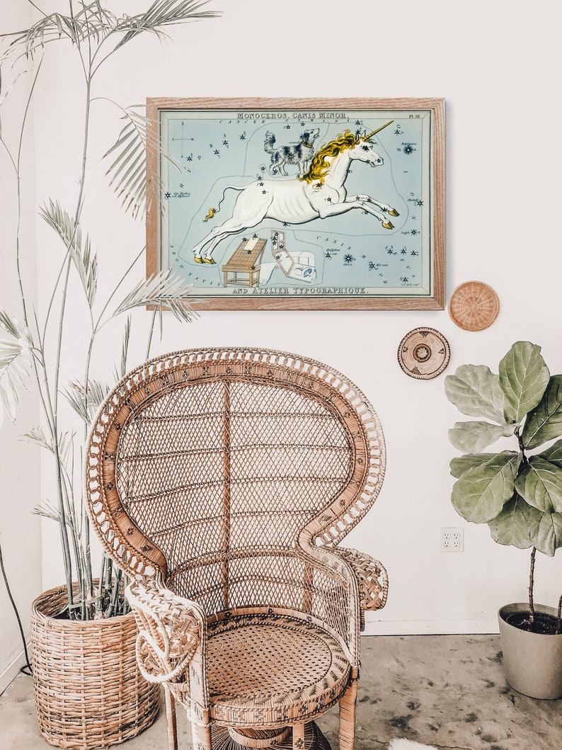 K&ouml;nnte beinhalten: Eine Vintage-Illustration des Sternbilds Einhorn, mit einem wei&szlig;en Einhorn mit goldenem Horn, vor einem blauen Hintergrund mit wei&szlig;en Sternen. Die Illustration ist in einem hellen Holzrahmen gerahmt und h&auml;ngt an einer wei&szlig;en Wand.