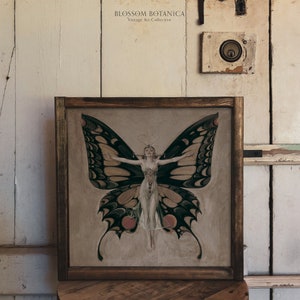 May include: A framed vintage art print featuring a woman with butterfly wings. The woman is wearing a white dress and has her arms outstretched. The butterfly wings are black, green, and yellow.