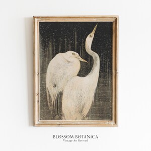 May include: A vintage framed print of two white birds with long necks and beaks, standing on a dark background with a textured pattern. The print is in a light wood frame.