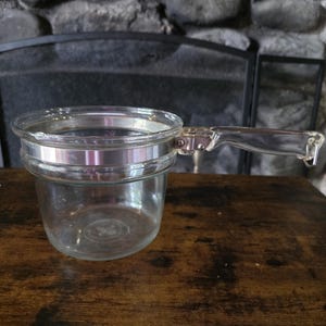 Könnte beinhalten: Ein klarer Glas-Stieltopf mit einem breiten, kreisförmigen Rand und einem transparenten Glasgriff, der mit einem silberfarbenen Metallband befestigt ist. Das Gefäß hat einen tiefen, zylindrischen Körper mit einem glatten, abgerundeten Boden.