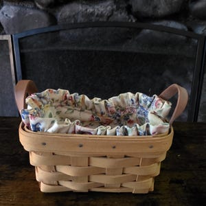 May include: A rectangular woven wood basket with two light brown leather handles. The basket features a removable fabric liner with a ruffled edge, decorated with a colourful floral pattern in shades of pink, blue, green, and yellow on a cream background.