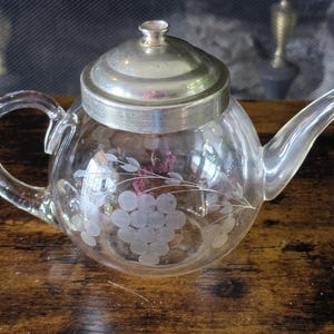 May include: A clear glass teapot with a silver lid and handle. The teapot has an etched grape design. It is on a wooden surface.