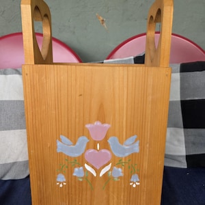 Op de afbeelding: Een rechthoekige houten tijdschriftenhouder met een lichtbruine afwerking en twee rechtopstaande handvatten met hartvormige uitsparingen. Het voorpaneel is versierd met een geschilderd ontwerp van twee lichtblauwe vogels, een roze hart, een roze tulp en blauwe bloemen.