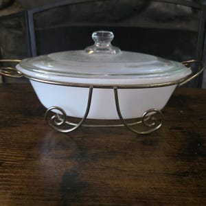 Puede incluir: Una fuente ovalada para horno hecha de vidrio opalino blanco con una tapa de vidrio transparente. La fuente se sostiene dentro de un soporte de alambre metálico dorado con asas y pies de volutas decorativas, elevando el recipiente.