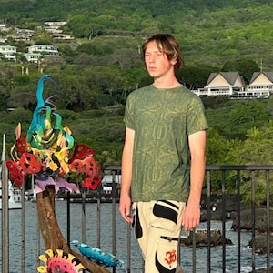 May include: A person wearing a green short-sleeved shirt with a geometric pattern and beige cargo shorts with a black and white design and an orange Om symbol. The person is standing on a concrete surface with a colorful metal sculpture in the background.